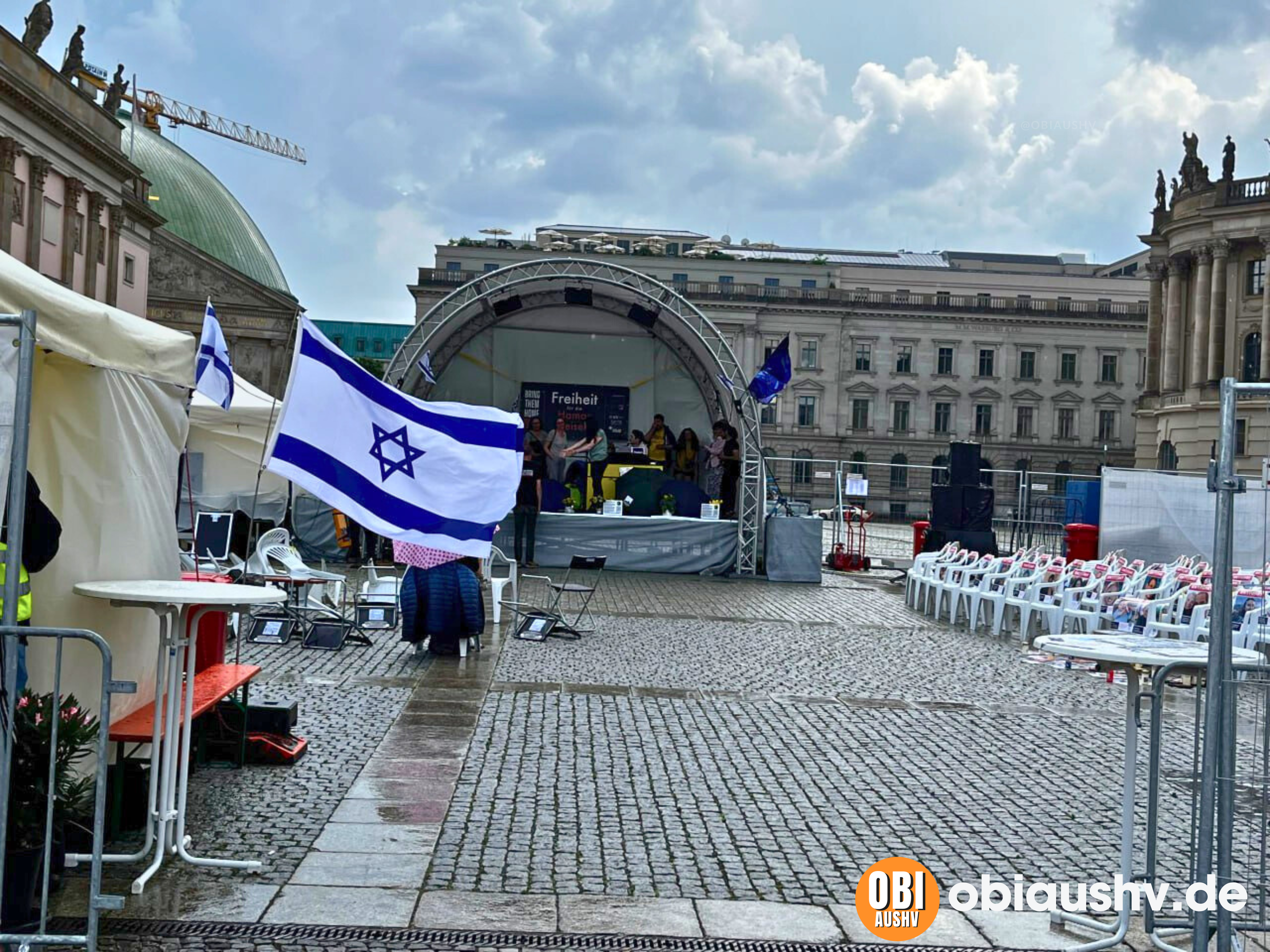Das Foto zeigt eine Außenszene mit einer Bühne und einem Sitzbereich. Auf der Bühne hängt ein Banner mit dem Wort “Freiheit”. Im Vordergrund sind zwei Flaggen von Israel deutlich zu sehen.