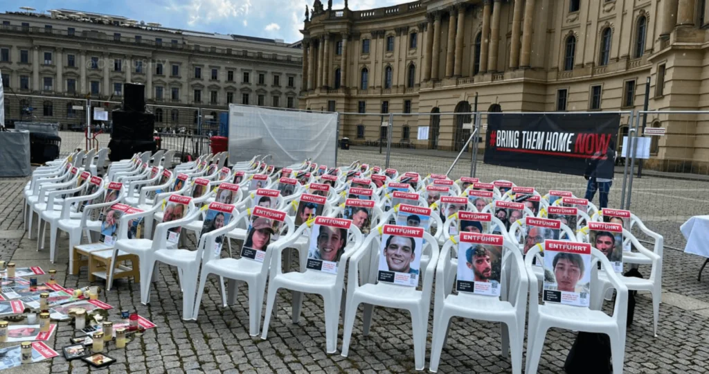 Hamas-Geiseln Israel Berlin Bebelplatz - obiaushv.de Das Foto zeigt eine Außenszene mit Reihen von weißen Plastikstühlen, die auf ein Banner gerichtet sind, auf dem “BRING THEM HOME NOW!” steht. An jedem Stuhl ist ein Foto befestigt, und es liegen verschiedene Gegenstände auf dem Boden vor den Stühlen, wie Bücher und persönliche Gegenstände. Im Hintergrund ist ein großes Gebäude mit klassischer Architektur zu sehen.