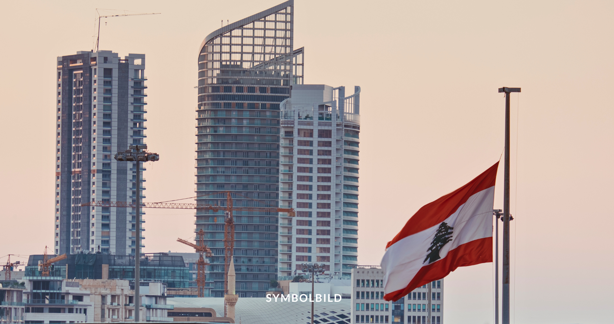 Das Bild zeigt eine Stadtsilhouette während der Dämmerung oder des Sonnenuntergangs. Mehrere im Bau befindliche Hochhäuser sind zu sehen, mit Kränen, die sich gegen den Himmel abheben. Im Vordergrund befindet sich ein Fahnenmast mit einer Libanon-Flagge. Symbolbild
