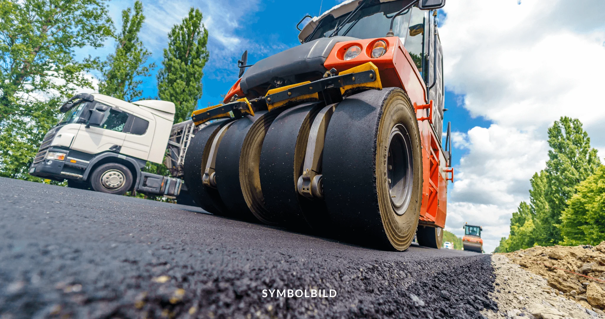 Das Bild zeigt eine Nahaufnahme eines großen orangefarbenen Straßenwalzen mit schwarzen Rädern auf einer Asphaltfläche. Im Hintergrund ist ein weißer Lastwagen teilweise sichtbar. Der Himmel ist teilweise bewölkt, und die Perspektive verleiht der Straßenwalze eine dramatische Wirkung, wodurch ihre Größe und ihre Rolle bei Straßenbauarbeiten oder -wartung betont werden. Der Text “SYMBOLBILD” ist unten im Bild eingeblendet, was darauf hinweist, dass dieses Bild repräsentativ oder illustrativ für etwas ist und keine tatsächliche Ereignis darstellt. A59 Vollsperrungen Symbolbild