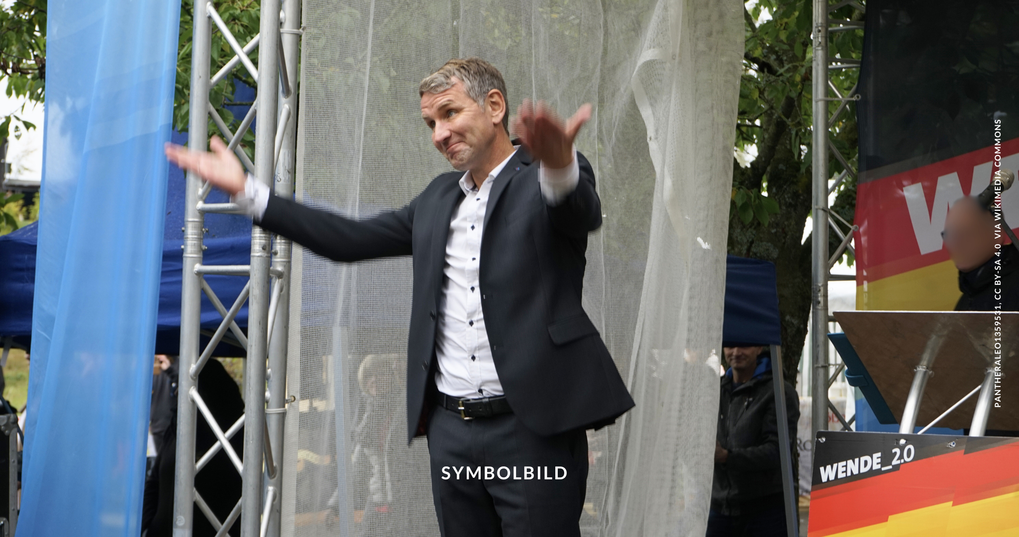 Das Bild zeigt Björn Höcke, der auf einer Bühne steht und die Arme ausgestreckt hat. Im Hintergrund sind Teile von Bannern oder Schildern zu sehen. Björn Höcke verurteilt Symbolbild
