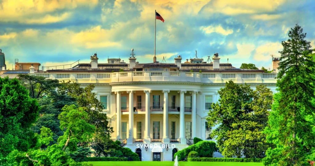 Das Bild zeigt das Weiße Haus, den offiziellen Wohn- und Arbeitsplatz des Präsidenten der Vereinigten Staaten, in Washington D.C. Die Aufnahme zeigt die ikonische neoklassizistische Fassade des Gebäudes mit Säulen und zahlreichen Fenstern. Auf dem Dach weht die amerikanische Flagge. Der Himmel im Hintergrund ist teilweise bewölkt und hat einen warmen Schimmer, was auf Sonnenaufgang oder Sonnenuntergang hindeutet. Joe Biden Corona Symbolbild