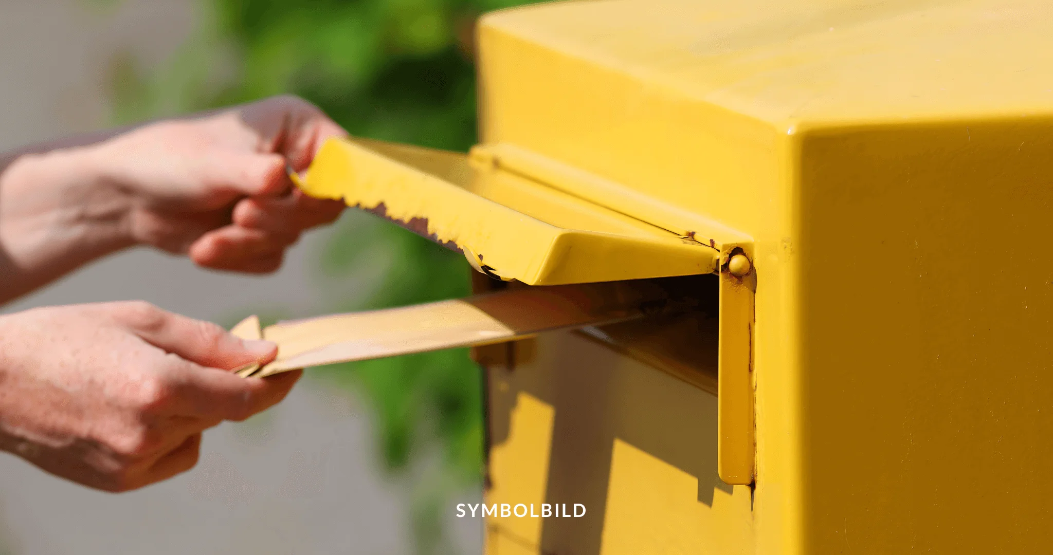 Das Bild zeigt eine Nahaufnahme von zwei Händen, die mit einem gelben Briefkasten interagieren. Eine Hand steckt einen Umschlag in den geöffneten Schlitz des Briefkastens. Postrecht Symbolbild