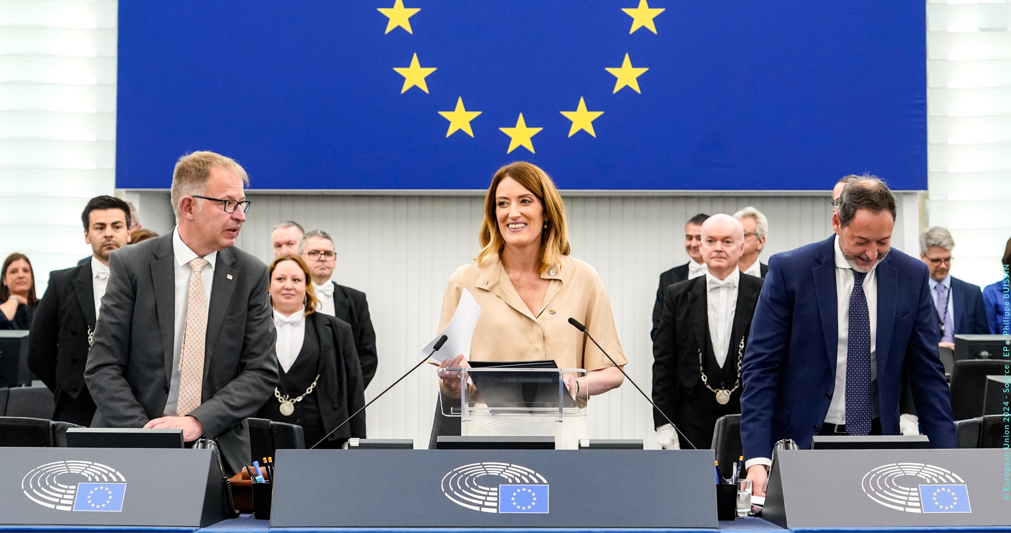 Das Bild zeigt eine Gruppe von Personen in einem formellen Rahmen im Europäischen Parlament. Im Mittelpunkt steht Roberta Metsola, die an einem Rednerpult mit Mikrofonen steht. Im Hintergrund ist die Flagge der Europäischen Union mit einem Kreis aus Sternen auf blauem Grund zu sehen.