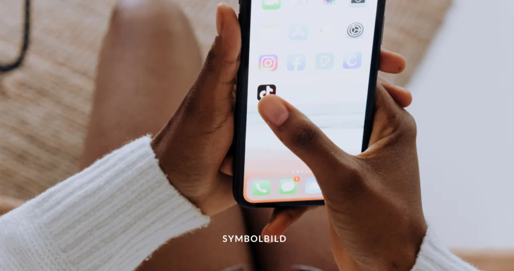 Das Bild wird analysiert: Gesichter sind möglicherweise verschwommen, um die Privatsphäre zu schützen. Gesendet von Copilot: Das Bild zeigt eine Nahaufnahme von zwei Händen, die ein Smartphone halten. Der Bildschirm des Smartphones ist sichtbar und zeigt verschiedene App-Symbole. Der Daumen zeigt auf die App TikTok. Symbolbild