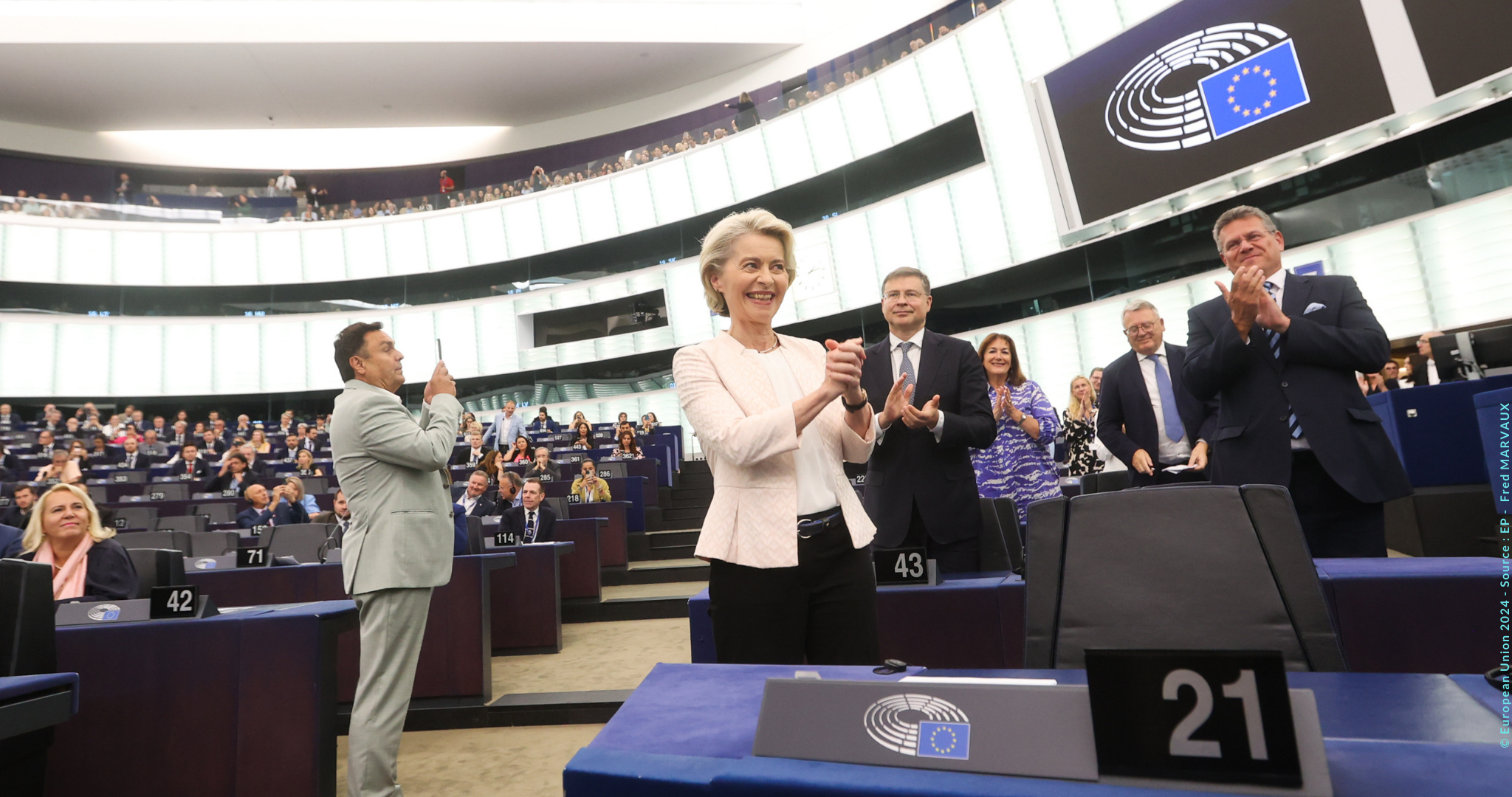 Das Bild zeigt eine Szene im Europäischen Parlament. Im Vordergrund steht Ursula von der Leyen. Um sie herum stehen mehrere Personen, die ebenfalls stehen und klatschen. Es zeigt den Moment nach der Wiederwahl.