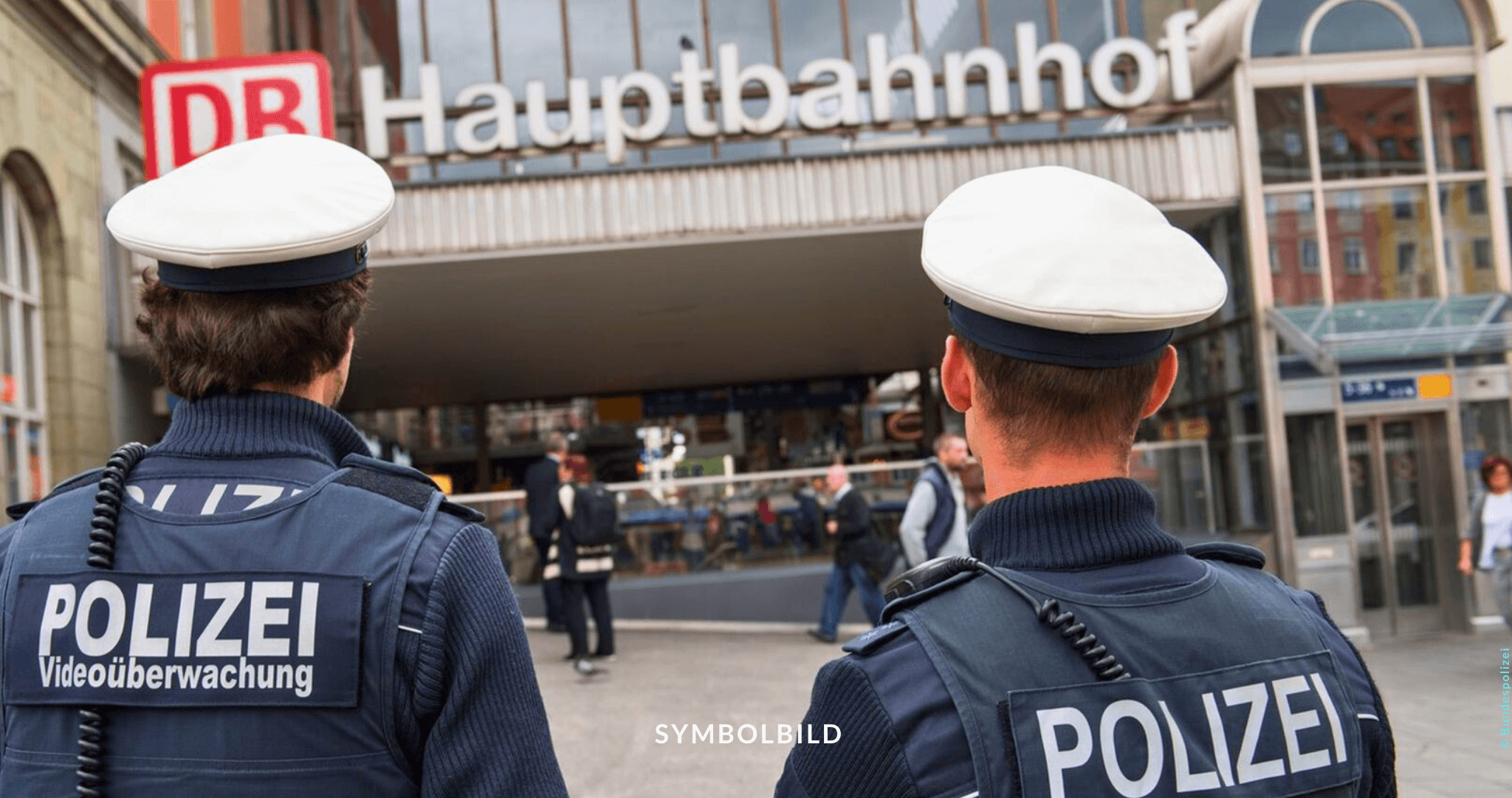 Das Bild zeigt zwei Polizisten von hinten, die auf ein Gebäude mit dem Schild “DB Hauptbahnhof” blicken. Die Polizisten tragen Uniformen mit der Aufschrift “Polizei” auf dem Rücken, sowie ein zusätzliches Etikett mit der Aufschrift “Videoüberwachung.” Gewalt Regionalbahn Symbolbild