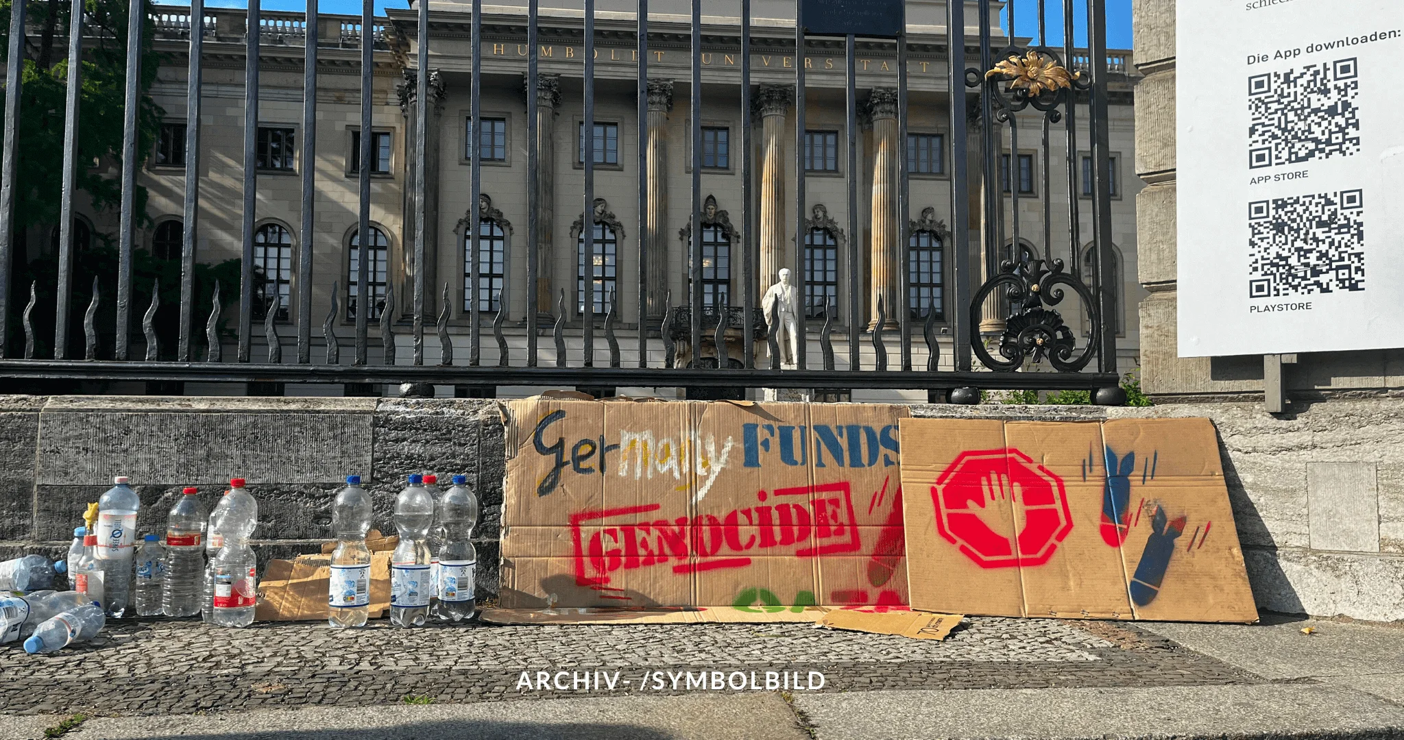 Das Bild zeigt ein provisorisches Pappschild, das vor einem Zaun aufgestellt ist. Auf dem Schild steht in gesprühter Schrift „Germany Funds Genocide“ („Deutschland finanziert Völkermord“). Neben dem Schild sind mehrere Plastikflaschen aufgereiht. Hinter dem Schild befindet sich ein Gebäude (Humboldt-Universität) mit klassischer Architektur und Statuen. Archiv-/Symbolbild DJV Gewalt gegen Journalisten