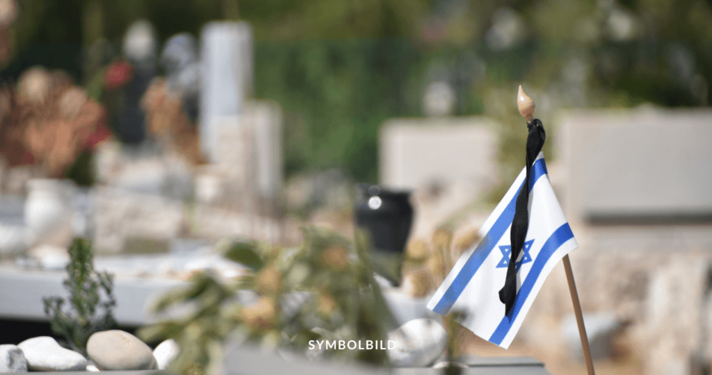 Das Bild zeigt eine kleine israelische Flagge, die an einem Stock befestigt ist. Die Flagge hat das charakteristische blaue Davidstern-Symbol zwischen zwei horizontalen blauen Streifen auf weißem Hintergrund. Im Hintergrund, der unscharf ist, sieht man einen Friedhof mit Grabsteinen und Blumengestecken. Am unteren Rand des Bildes steht das Wort “SYMBOLBILD”, was darauf hinweist, dass dieses Bild repräsentativ oder illustrativ für etwas ist. Israel: Fünf Geiseln tot aus Gaza geborgen