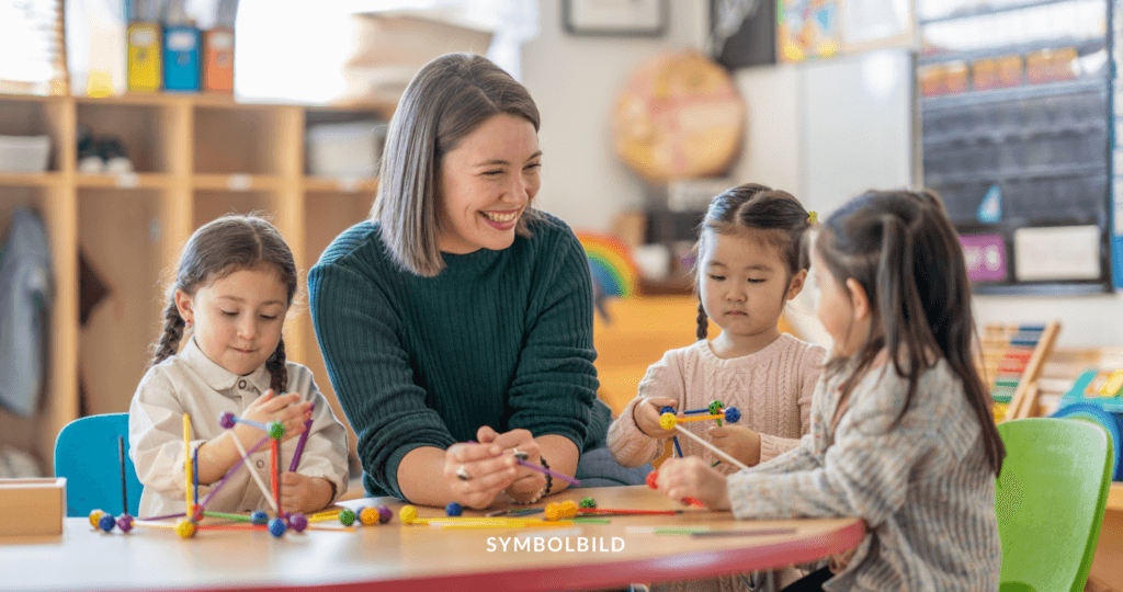 Das Bild zeigt vier Kinder und eine Frau, die an einem Tisch sitzen. Die Kinder hantieren mit verschiedenen Gegenständen. Das Bild zeigt eine typische Kita-Szene. Am unteren Rand des Bildes steht das Wort “SYMBOLBILD”. Kita Ausfallzeiten