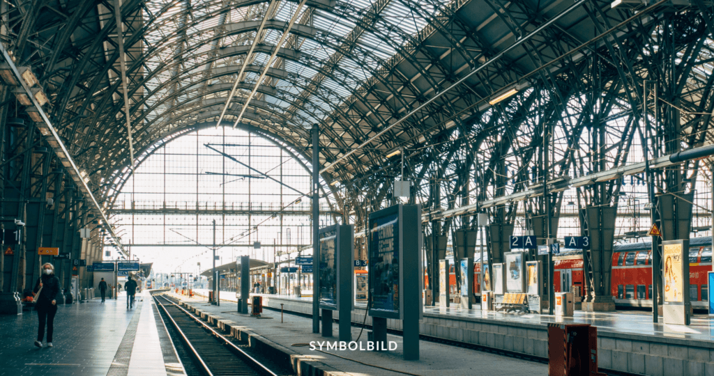 Das Bild zeigt eine Innenansicht des Hauptbahnhofes Frankfurt mit einem großen, gewölbten Glas- und Stahl-Dach. Mehrere Gleise führen zu Bahnsteigen, die mit Schildern wie “2A” und “3” gekennzeichnet sind. Auf den Bahnsteigen sind einige Personen zu sehen, und es sind Züge im Bahnhof geparkt. Schüsse Frankfurt Symbolbild