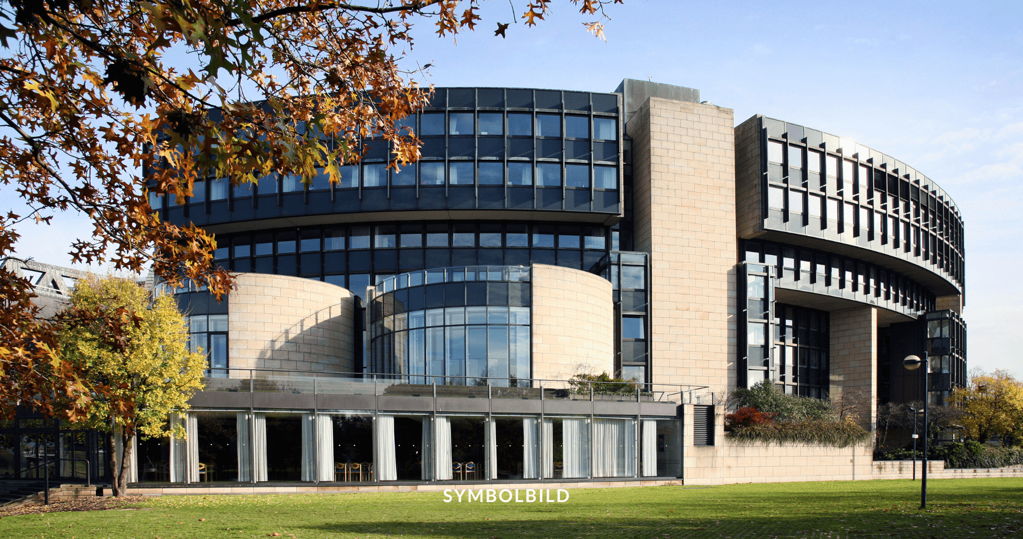 Das Bild zeigt ein modernes Gebäude (Landtag Düsseldorf) mit einer geschwungenen Architektur, das aus einer Mischung aus Glas- und Betonelementen besteht. Das Gebäude hat mehrere Ebenen mit reflektierenden Fenstern und ist von Grünflächen umgeben, darunter Bäume mit herbstlich gefärbten Blättern. Symbolbild Solingen Anschlag