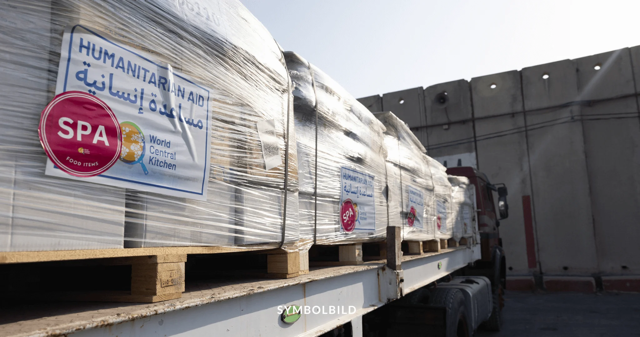 Das Bild zeigt einen großen Lastwagen, der mit Paletten beladen ist. Diese Paletten sind in durchsichtiger Plastikfolie eingewickelt und tragen mehrere Logos und Texte, die auf humanitäre Hilfe hinweisen. Zu den Logos gehören “World Central Kitchen”. Darüber steht der Text “HUMANITARIAN AID”. Im Hintergrund sieht man die hohe Mauer des Grenzübergangs Kerem Shalom. UNRWA Symbolbild