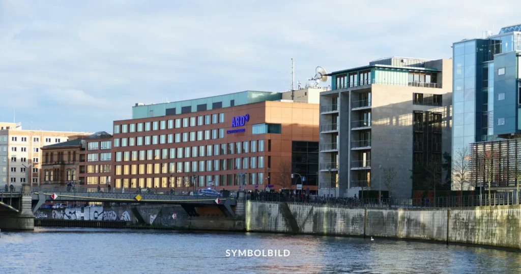 Das Bild zeigt das Hauptstadtstudio der ARD, eine Gemeinschaftseinrichtung der deutschen öffentlich-rechtlichen Rundfunkanstalten. Das Gebäude hat eine moderne Architektur mit mehreren Stockwerken und großen Fenstern. Es befindet sich neben einem Gewässer mit einer Betonböschung. Im Vordergrund ist eine Wand mit Graffiti zu sehen. Der Himmel ist teilweise bewölkt. SYMBOLBILD Streiks bei ARD und ZDF führen zu Programmstörungen und Tarifverhandlungen.