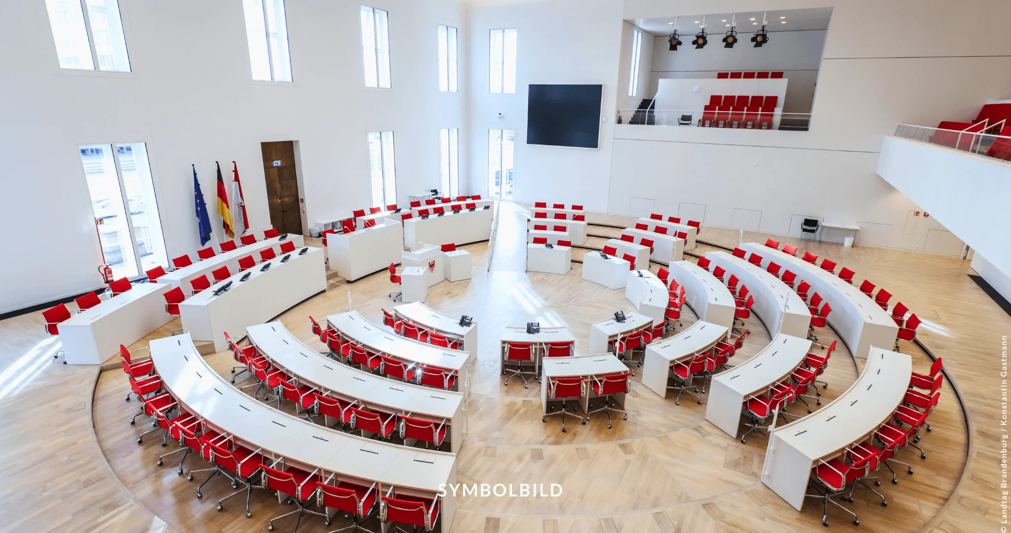 Das Bild zeigt den Plenarsaal des Landtags Brandenburg. Der Saal hat eine halbkreisförmige Anordnung von Tischen und roten Stühlen. Der Raum ist geräumig, mit hohen Decken und großen Fenstern, die viel natürliches Licht hereinlassen. Über dem Hauptbereich befindet sich eine Galerie für Besucher. Die Wände sind weiß, was einen Kontrast zu den roten Sitzmöbeln bildet, und an einer Seite des Raumes ist eine Flagge zu sehen. SYMBOLDBILD Landtagswahl in Brandenburg: SPD und AfD als stärkste Parteien bei komplexer Regierungsbildung.