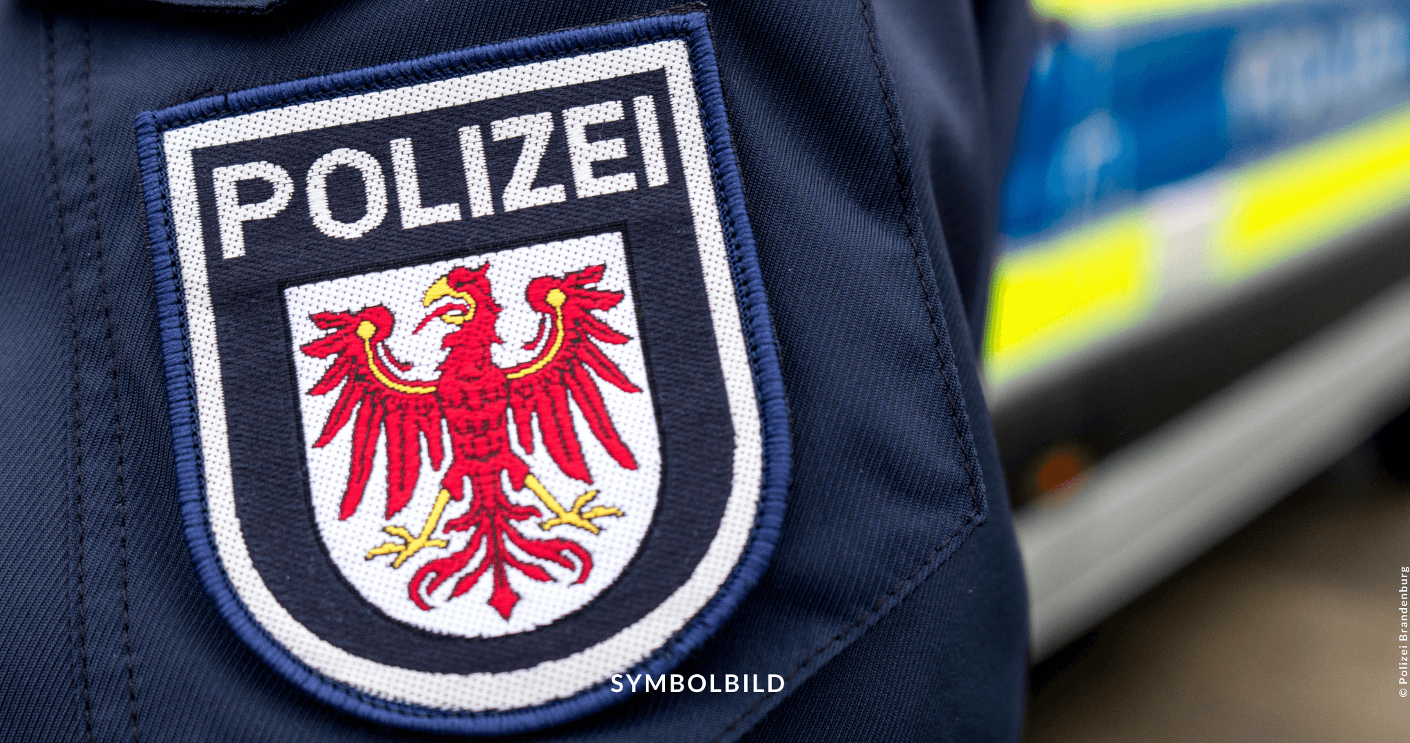 Das Bild zeigt ein Nahaufnahme eines Stoffabzeichens mit dem Wort “POLIZEI” in weißen Buchstaben auf dunklem Hintergrund. Darüber befindet sich ein Wappen mit einem roten Adler, der detailliert dargestellt ist und in einem schildförmigen Rahmen erscheint. Im Hintergrund ist unscharf ein Fahrzeug mit reflektierenden gelben und blauen Streifen zu erkennen, was typisch für Polizeifahrzeuge ist. SYMBOLBILD für das Verbot Islamisches Zentrum Fürstenwalde vom Innenministerium Brandenburg.