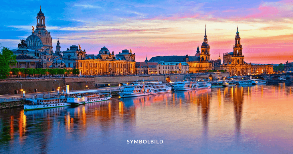 Das Bild zeigt eine Panoramaansicht von Dresden bei Dämmerung. Der Himmel ist in warmen Orangetönen und blau gefärbt. Die Architektur wirkt europäisch, mit historischen Gebäuden, die Kuppeln und Türme aufweisen. Die Elbe (Fluss) fließt durch die Stadt und spiegelt die Gebäude und den Himmel wider. Am Flussufer sind Boote zu sehen. SYMBOLBILD Einschleusungen in Dresden