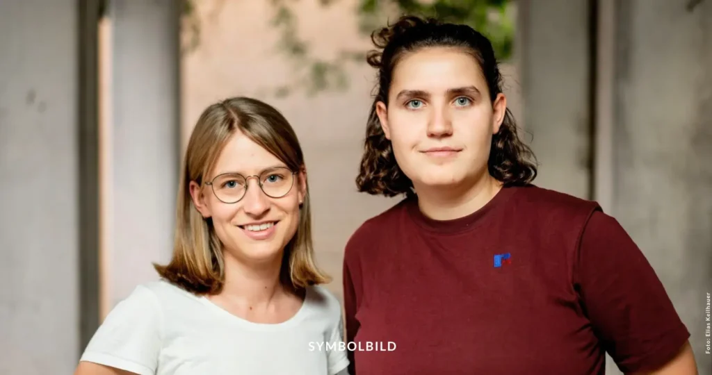 Das Bild zeigt zwei Personen, die nebeneinander stehen (Svenja Appuhn und Katharina Stolla, die Bundessprecher*innen Grüne Jugend). Die Person links trägt ein weißes Oberteil, während die Person rechts ein rotes Oberteil mit einem blauen Logo auf der linken Brustseite trägt. Der Hintergrund ist unscharf, deutet aber auf eine Innenaufnahme mit etwas Grün im Hintergrund hin. SYMBOLBILD Rücktritt des Bundesvorstands der Grünen Jugend aus Enttäuschung über Parteipolitik