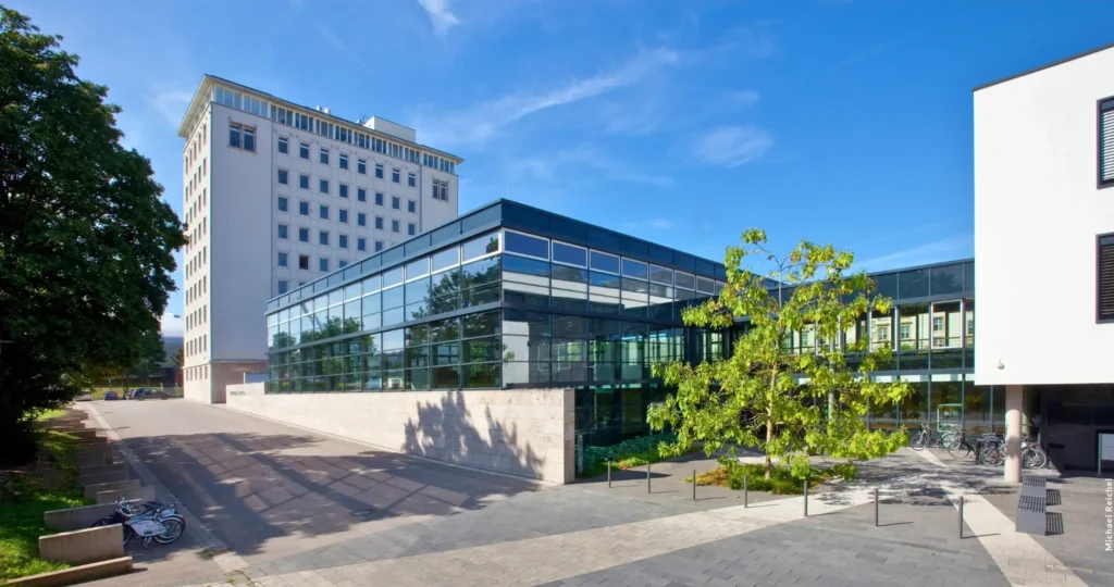 Das Bild zeigt den Thüringer Landtag, ein modernes Gebäude mit einer Kombination aus Glas- und Betonstrukturen. Im Vordergrund befindet sich ein offener Platz mit ordentlich angeordneten Bänken und einigen Bäumen, die Schatten spenden. Symbolbild AfD-Verbot nach chaotischer Sitzung durch Innenminister Georg Maier gefordert