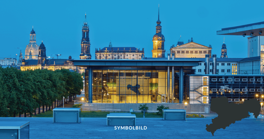 Im Vordergrund befindet sich ein modernes Glasgebäude mit eingeschaltetem Licht, das die Silhouette einer Reiterstatue zeigt. Dahinter sind historische Gebäude mit Türmen und aufwendiger Architektur zu sehen, die gegen den Abendhimmel beleuchtet sind. Außenansicht des Plenarsaals mit Blick auf die Dresdner Altstadt. SYMBOLBILD Sachsen Landtagswahl