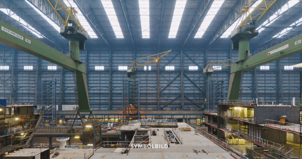 Das Bild zeigt einen großen industriellen Innenraum, der Meyer Werft, mit hohen Decken und großen Fenstern, die natürliches Licht hereinlassen. Es zeigt zwei grüne Portalkräne (auf Schien), und darunter ist ein aktiver Bau- oder Fertigungsbereich mit verschiedenen Strukturen und Ausrüstungen. Symbolbild zeigt Meyer Werft, Bundestag