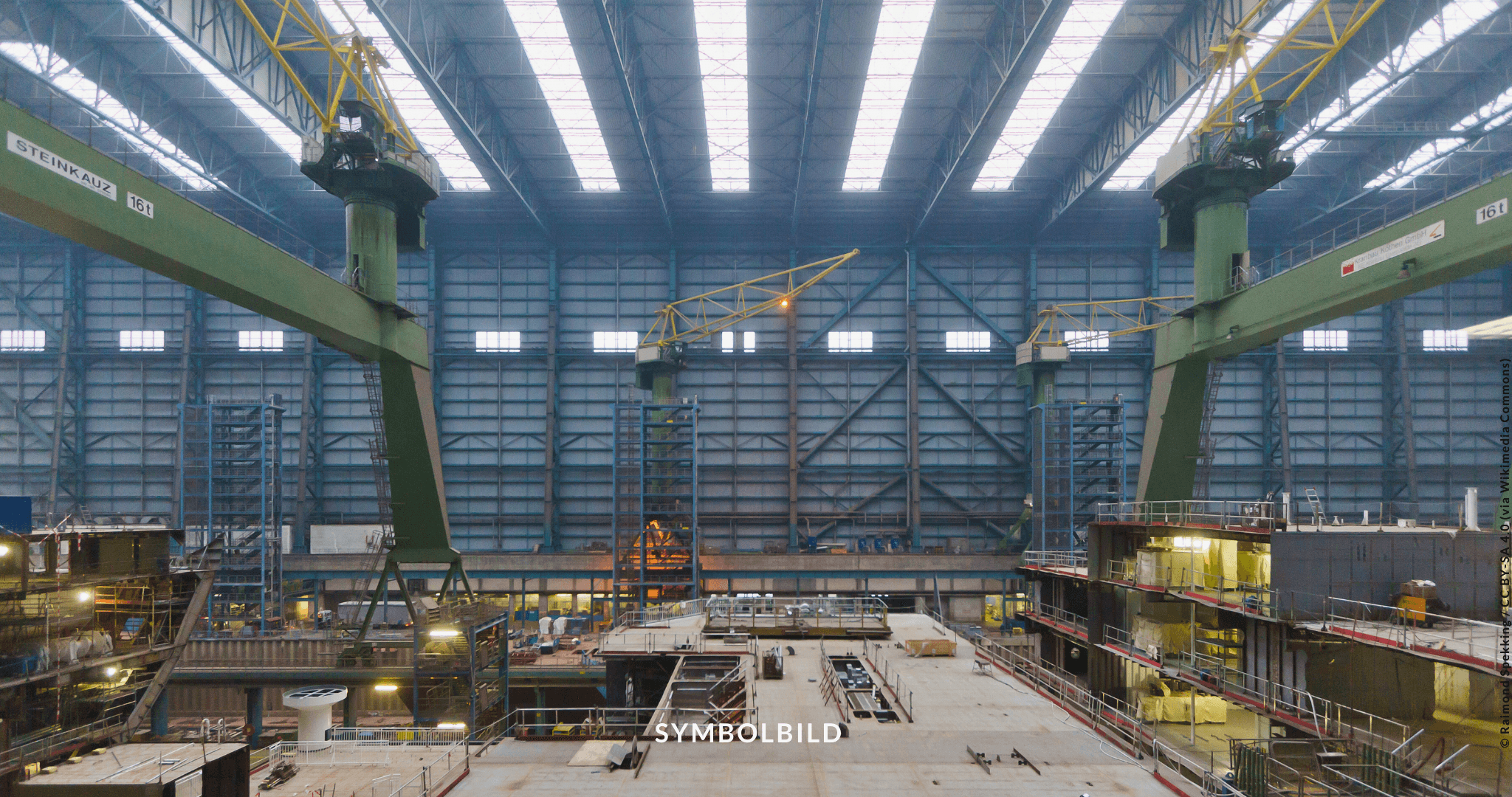 Das Bild zeigt einen großen industriellen Innenraum, der Meyer Werft, mit hohen Decken und großen Fenstern, die natürliches Licht hereinlassen. Es zeigt zwei grüne Portalkräne (auf Schien), und darunter ist ein aktiver Bau- oder Fertigungsbereich mit verschiedenen Strukturen und Ausrüstungen. Symbolbild zeigt Meyer Werft, Bundestag