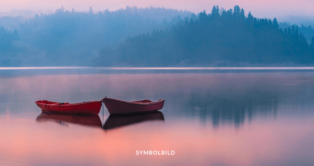 Das Bild zeigt zwei Boote, eines rot und eines grau, die auf einem ruhigen Gewässer schwimmen. Die Atmosphäre ist neblig, und im Hintergrund sind die Silhouetten bewaldeter Hügel oder Berge zu sehen. Die Spiegelung der Boote und der Hügel ist auf der Wasseroberfläche sichtbar, was eine friedliche und ruhige Szene schafft. Symbolbild für den Anstieg der Suizidrate in Deutschland.