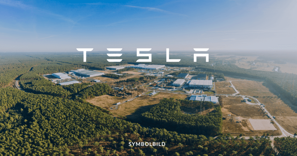 Das Bild zeigt eine Luftaufnahme der Tesla-Fabrik in Grünheide, Deutschland. Die Fabrikgebäude sind groß und weitläufig, mit mehreren Abschnitten und Dächern. Die umliegende Gegend ist stark bewaldet, mit Bäumen, die sich bis zum Horizont erstrecken, außer dort, wo eine Straße die Landschaft durchschneidet. Im oberen Teil des Bildes ist das Tesla-Logo prominent zu sehen. SYMBOLBILD Tesla Hausbesuche bei krankgeschriebenen Mitarbeitern in Grünheide