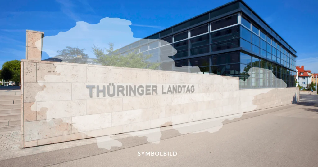 Das Bild zeigt ein modernes Gebäude mit großen Glasfenstern und einem klaren blauen Himmel darüber. Vor dem Gebäude befindet sich eine Mauer mit der Aufschrift “THÜRINGER LANDTAG”. Über dem Bild liegt die Umrisskarte von Thüringen. SYMBOLBILD Thüringen, Wahlen 2024, Landtagswahl