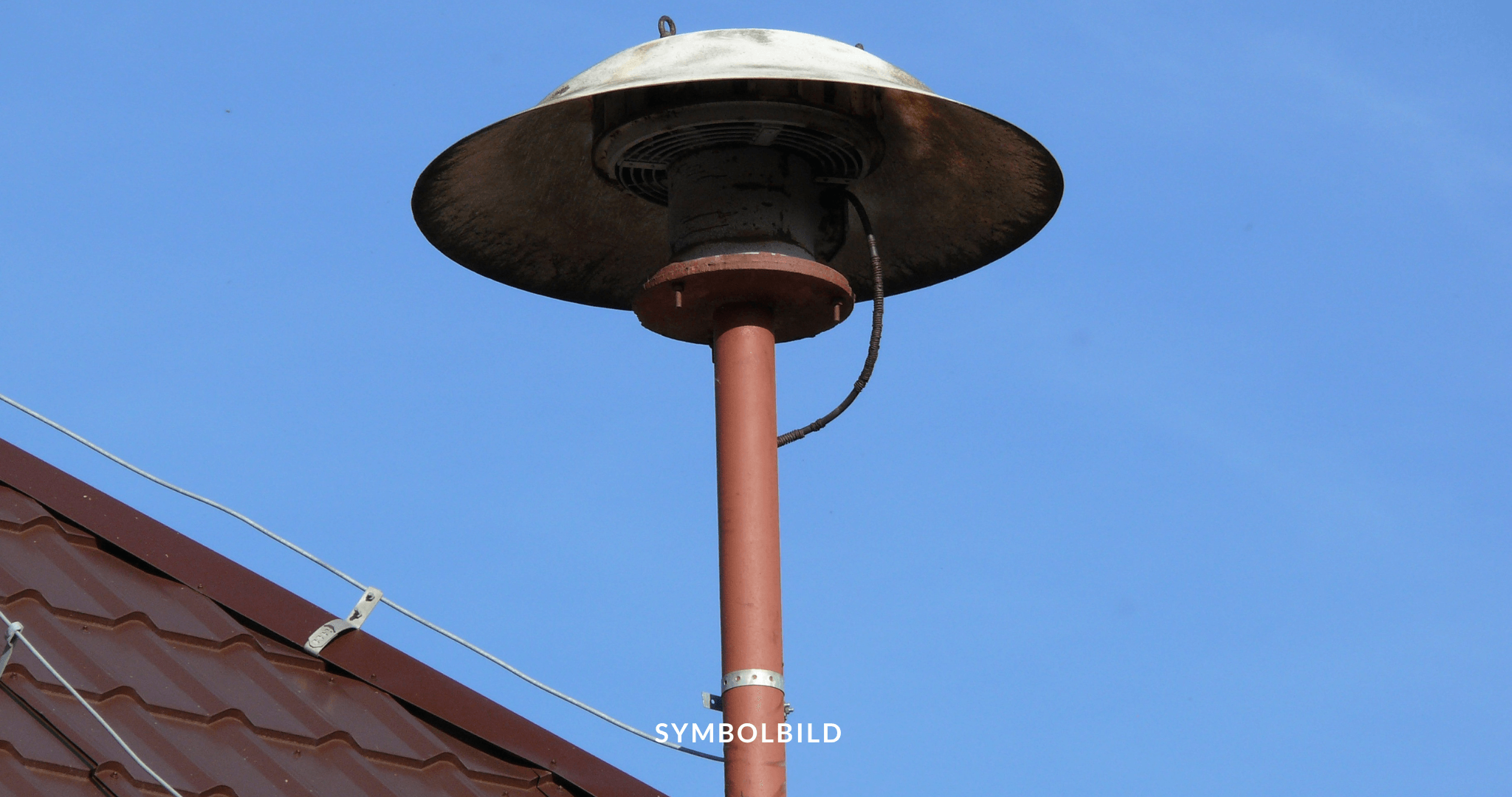 Das Bild zeigt eine Außensirene, die an einem Mast auf einem schrägen Dach montiert ist. Vor einem himmelblauen Hintergrund ist am unteren Rand das Build mit dem Wort “SYMBOLBILD” überlagert. Symbolbild für Warntag