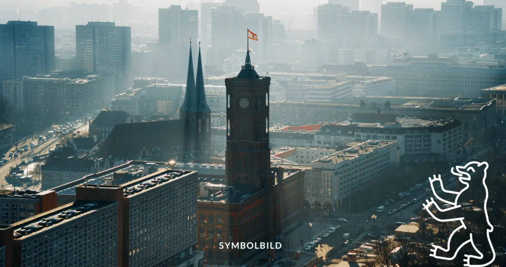 Das Bild zeigt eine Luftaufnahme von Berlin mit einem markanten Gebäude, das einen großen Uhrturm mit einer Fahne an der Spitze aufweist. Dieses Gebäude ist das Rote Rathaus in Berlin. Die umliegende Gegend umfasst verschiedene andere Gebäude mit unterschiedlichen architektonischen Stilen und Größen, Straßen mit sichtbarem Verkehr und im Hintergrund eine Kirche mit zwei Türmen. Symbolbild Kita-Streik in Berlin: Mitarbeiter fordern bessere Arbeitsbedingungen für städtische Kitas.