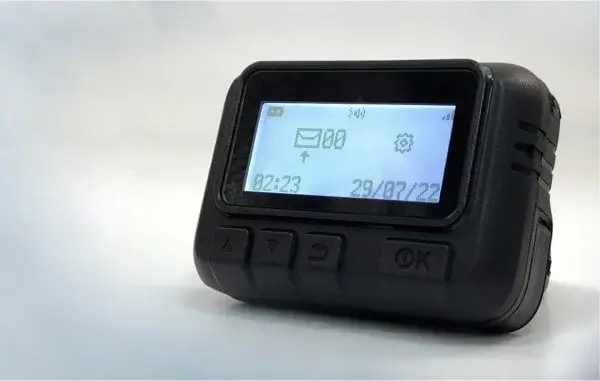 Das Bild zeigt einen schwarzen Pager, speziell das Modell Gold Apollo AR-924. Das Gerät hat einen kleinen Bildschirm, der verschiedene Symbole und Informationen anzeigt: ein Batterieanzeigesymbol oben links, daneben die Signalstärke und möglicherweise ein Umschlagsymbol, das auf neue Nachrichten oder Benachrichtigungen hinweist. Darunter wird die Uhrzeit “02:23” und das Datum “29/07/22” angezeigt. Am unteren Rand des Geräts befinden sich vier Tasten, die von links nach rechts mit einem Ein-/Ausschalt-Symbol, zwei Pfeilen nach oben und unten sowie einer “OK”-Taste beschriftet sind. Der Hintergrund ist schlicht, wodurch der Pager selbst hervorgehoben wird.