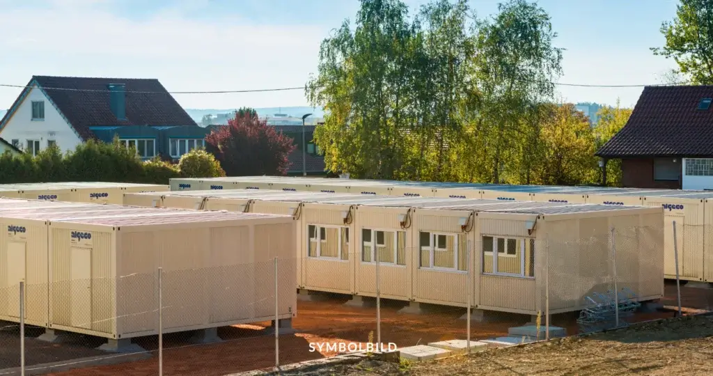 Das Bild zeigt mehrere Containergebäude, die als temporäre Unterkünfte für Geflüchtete dienen. Die Container sind in Reihen aufgestellt und durch einen Zaun mit Maschendraht abgesichert. Im Hintergrund sind Wohnhäuser und Bäume zu sehen. Symbolbild Geflüchtete erhalten 2025 weniger finanzielle Unterstützung durch Kürzung der Bedarfssätze.