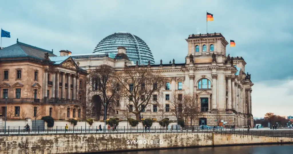 Das Bild zeigt das Reichstagsgebäude in Berlin, Deutschland. Es ist ein historisches Gebäude, das als Sitz des Deutschen Bundestages dient. Auffällig ist die große Glaskuppel auf dem Dach des Gebäudes, die von Sir Norman Foster entworfen wurde. Auf dem Gebäude wehen zwei deutsche Flaggen, und links ist eine Flagge der Europäischen Union zu sehen. Im Vordergrund verläuft ein Fluss, und entlang des Ufers sind einige Menschen zu Fuß unterwegs. SYMBOLBILD Sicherheitspaket der Bundesregierung Gesetzesverschärfungen vor finaler Abstimmung im Bundestag