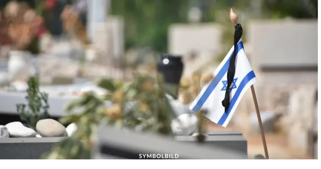 Das Bild zeigt eine kleine israelische Flagge, die an einem dünnen Stab befestigt ist. Die Flagge hat das charakteristische blaue Davidstern-Symbol zwischen zwei horizontalen blauen Streifen auf weißem Hintergrund. Im Hintergrund, der unscharf ist, sieht man einen Friedhof mit Grabsteinen. Symbolbild Terrorangriff Israel 7. Oktober Gedenken an die Opfer und Geiseln des Massakers