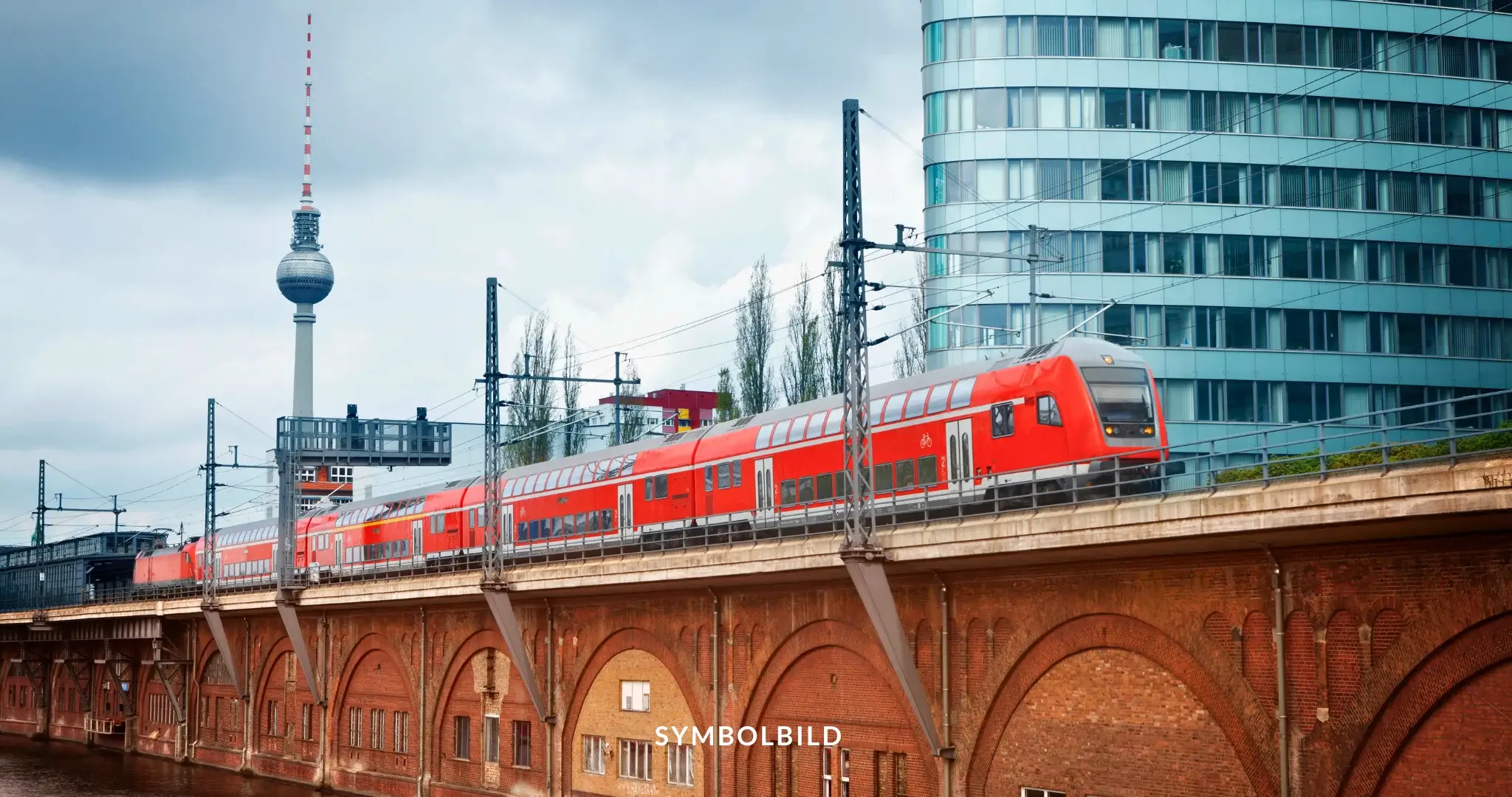 Das Bild zeigt einen roten Zug, der auf einer erhöhten Eisenbahnstrecke fährt. Im Hintergrund ragt der Berliner Fernsehturm empor, ein markantes Wahrzeichen der Stadt. Der Zug passiert ein modernes, rundes Gebäude mit vielen Fenstern. Unter der Eisenbahnstrecke sieht man eine Backsteinmauer mit Bögen. Der Himmel ist bewölkt. SYMBOLBILD Verkehrseinschränkungen in Berlin wegen Biden-Besuch, 17. und 18. Oktober 2024