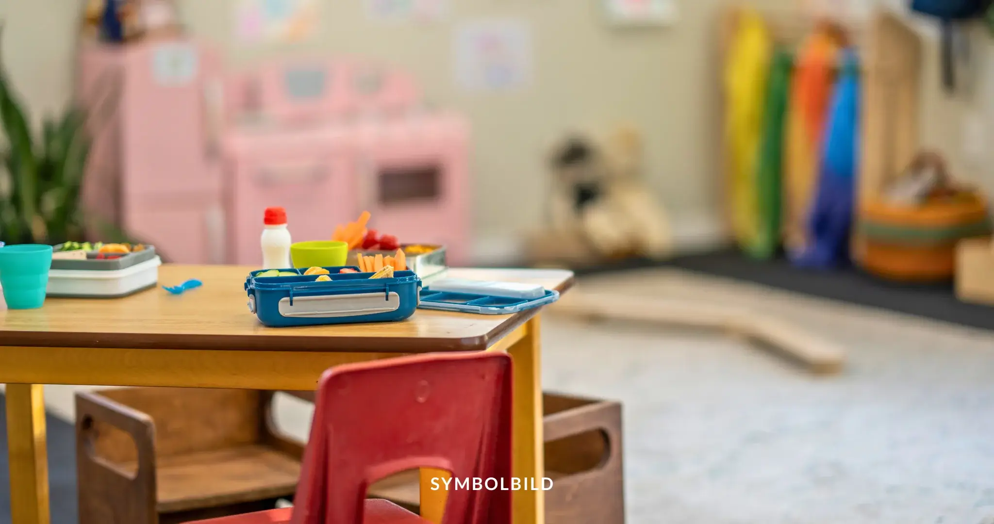 Das Bild zeigt eine bunte Kita-Umgebung mit einem Kindertisch im Vordergrund. Auf dem Tisch befinden sich verschiedene Gegenstände, darunter eine Brotdose und einige Plastikbehälter. Im Hintergrund sind Kindermöbel und Spielzeug unscharf zu erkennen, was auf einen Spielbereich hinweist. SYMBOLBILD Arbeitsgericht Berlin erklärt Streikverbot für kommunale Kitas, Verdi bereitet Berufung vor