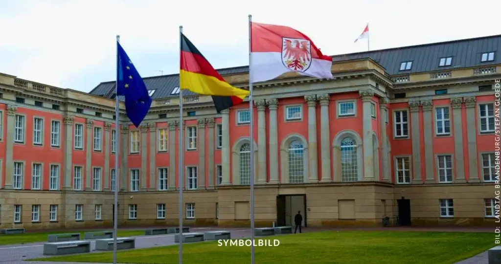 Das Bild zeigt den Innenhof mit Haupteingang des Landtagsgebäudes Brandenburg in Potsdam. Im Vordergrund sind drei Flaggenmasten zu sehen, an denen die Flaggen der Europäischen Union, Deutschlands und Brandenburgs wehen. Das Gebäude im Hintergrund hat eine auffällige Fassade in Rot- und Beigetönen mit großen Fenstern und Säulen. Auf der rechten Seite des Bildes ist der Text "BILD: LANDTAG BRANDENBURG/STEFAN GLOEDE" zu sehen. Am unteren Rand des Bildes steht das Wort "SYMBOLBILD." SYMBOLBILD Brandenburg: Koalitionsvertrag unterzeichnet