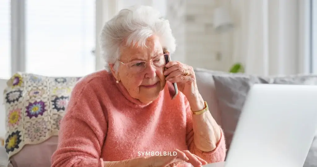 Das Bild zeigt eine ältere Person, die ein Smartphone ans Ohr hält und vor einem Laptop sitzt. Die Person trägt einen rosa Pullover und hat weißes Haar. Im Hintergrund ist eine gehäkelte Decke mit bunten Blumenmustern zu sehen. SYMBOLBILD Enkeltrick-Banden durch länderübergreifenden Polizeieinsatz zerschlagen