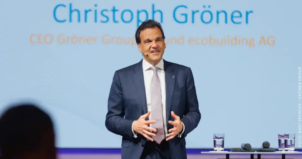 Das Bild zeigt Christoph Gröner, CEO der Gröner Group und ecobuilding AG. Er steht vor einer Leinwand, auf der sein Name und seine Titel in blauer und orangefarbener Schrift zu sehen sind. Christoph Gröner trägt einen dunklen Anzug mit weißem Hemd und einer hellen Krawatte. Vor ihm auf einem Tisch stehen zwei Gläser Wasser und zwei Mikrofone. Der Hintergrund ist hellblau gehalten. Bild: Gröner Gruppe wegen Insolvenzverschleppung und Sozialversicherungsbetrug.