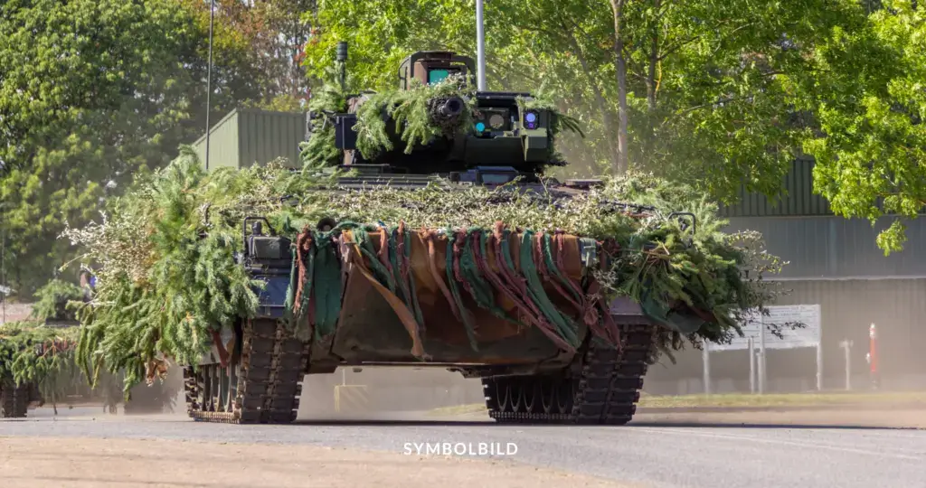 Das Bild zeigt ein gepanzertes Militärfahrzeug der Bundeswehr, das mit Tarnmaterial bedeckt ist. Das Fahrzeug ist mit Zweigen und Blättern bedeckt, um es in einer bewaldeten Umgebung zu tarnen. Es fährt auf einer Straße, und im Hintergrund sind Bäume und ein Gebäude zu sehen. Das Fahrzeug hat Kettenantrieb und ist mit verschiedenen Ausrüstungsgegenständen ausgestattet. Am unteren Rand des Bildes steht das Wort „SYMBOLBILD“. Bundeswehrmandate verlängert und neue Truppenstationierung in Litauen