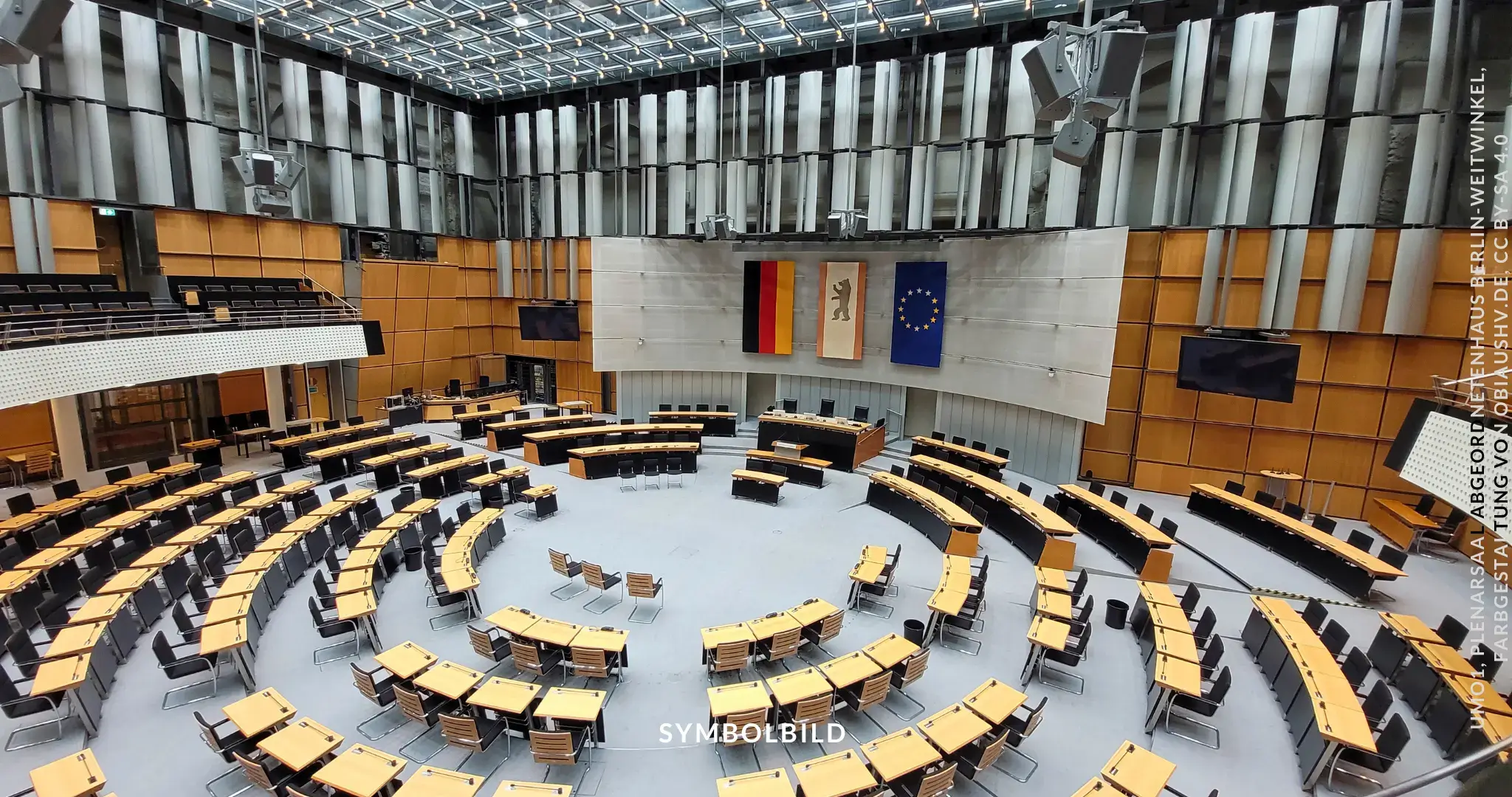 Das Bild zeigt den Plenarsaal des Abgeordnetenhauses von Berlin. Der Saal hat eine moderne Architektur mit einer großen Glasdecke, die viel Licht hereinlässt. Die Sitzanordnung ist halbkreisförmig und auf mehreren Ebenen angeordnet, was eine gute Sicht auf das Rednerpult und die zentrale Bühne ermöglicht. An der Wand hinter dem Rednerpult hängen drei Flaggen: die deutsche Nationalflagge, die Berliner Landesflagge und die Europaflagge. SYMBOLBILD Die Entscheidung zu Berlins Wiederholungswahl schließt das Kapitel der Berliner Pannenwahl