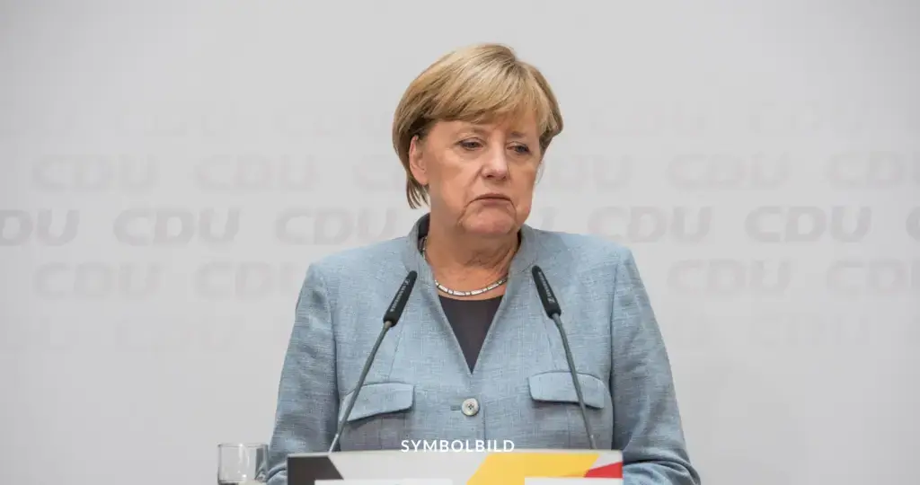 Das Bild zeigt Angela Merkel, die hinter einem Rednerpult mit zwei Mikrofonen steht. Im Hintergrund ist ein wiederholtes Muster der Buchstaben "CDU" zu sehen, was für "Christlich Demokratische Union" steht, eine bedeutende politische Partei in Deutschland. Angela Merkel trägt einen hellblauen Blazer und eine Halskette. Am unteren Rand des Bildes steht das Wort "SYMBOLBILD". Merkel kritisiert Merz für AfD-Zusammenarbeit