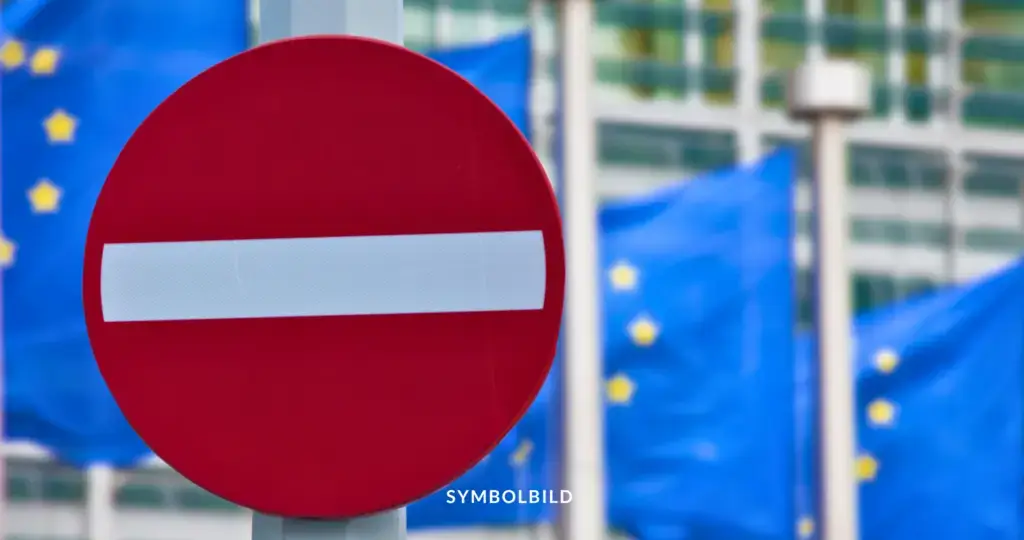 Das Bild zeigt ein Verkehrsschild mit einem roten Kreis und einem weißen horizontalen Balken in der Mitte, das ein Einfahrverbot signalisiert. Im Hintergrund sind mehrere blaue Flaggen mit gelben Sternen zu sehen, die die Flagge der Europäischen Union darstellen. Unter dem Schild steht das Wort "SYMBOLBILD". SYMBOLBILD Neue EU-Zölle auf russische Agrarimporte zur Schwächung der Wirtschaft.