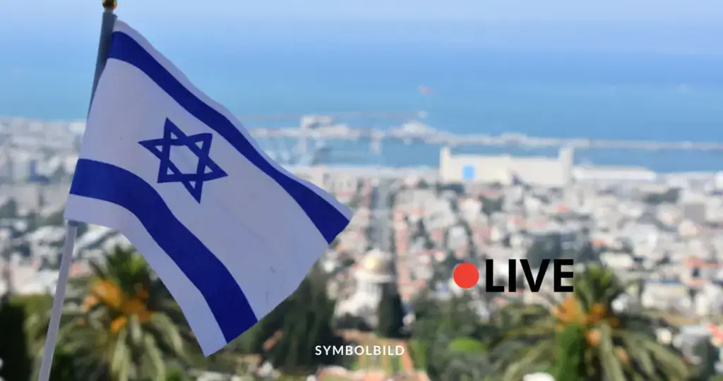 Das Bild zeigt die Flagge Israels im Vordergrund, mit einer Stadtansicht und dem Meer im Hintergrund. Die Flagge hat einen blauen Davidstern in der Mitte, flankiert von zwei horizontalen blauen Streifen oben und unten. Die Stadt im Hintergrund scheint Haifa zu sein, erkennbar an den Bahá'í-Gärten und dem Hafengebiet. Auf der rechten Seite des Bildes ist das Wort „LIVE“ mit einem roten Punkt daneben zu sehen.