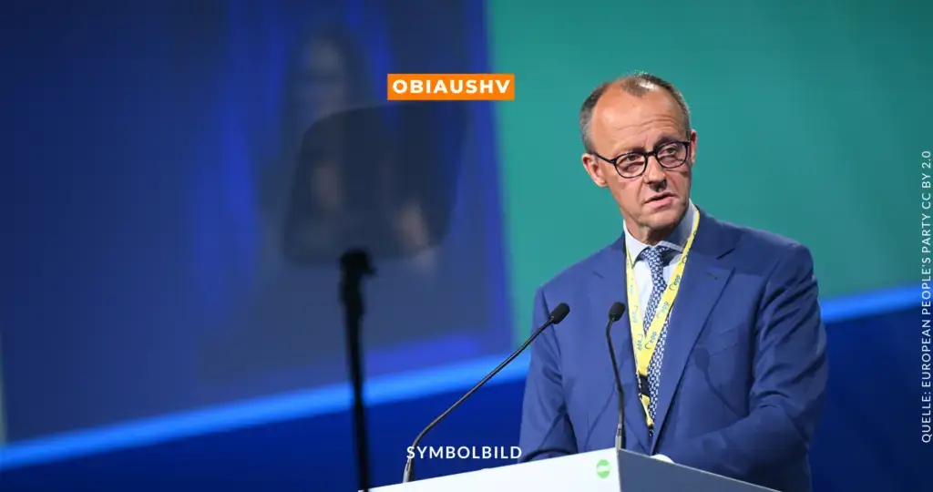 Das Bild zeigt Friedrich Merz, einen deutschen Politiker der CDU, während einer öffentlichen Rede. Er trägt einen blauen Anzug, ein weißes Hemd und eine gemusterte Krawatte. Ein gelbes Lanyard hängt um seinen Hals. Im Hintergrund ist ein bläulich-grüner Hintergrund zu sehen, und es gibt eine Leinwand mit einem Mikrofon vor ihm sowie einen Teleprompter auf der linken Seite. Oben in der Mitte des Bildes steht in einem orangefarbenen Kasten der Text „OBIAUSHV“, und unten in der Mitte steht in weißer Schrift „SYMBOLBILD“. Auf der rechten Seite des Bildes ist der Text „QUELLE: EUROPEAN PEOPLE'S PARTY CC BY 2.0“ zu sehen. Symbolbild CDU-Parteitag Berlin 2025 – Friedrich Merz und das Sofortprogramm zur Asylpolitik