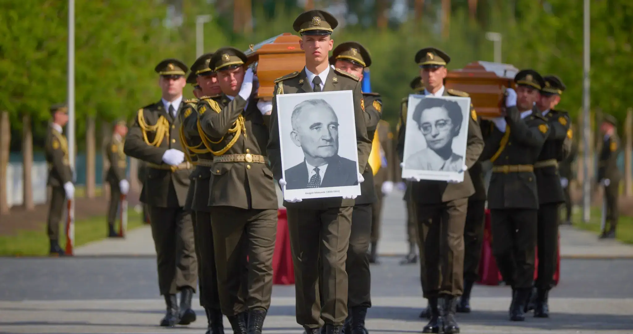 Mehrere ukrainische Soldaten in dunklen Paradeuniformen tragen bei einer militärischen Zeremonie zwei braune Holzsärge über eine breite Allee des Nationalen Militärfriedhofs nahe Kyjiw. Vor den Särgen marschieren zwei Soldaten, die große Schwarzweißporträts von Andrij Melnyk und seiner Ehefrau Sofiia Fedak-Melnyk halten. Melnyk ist als älterer Mann mit kurzem grauem Haar und Anzug dargestellt, seine Ehefrau mit Brille. Die Soldaten tragen Schirmmützen, weiße Handschuhe und goldene Schulterkordeln. Im Hintergrund stehen weitere uniformierte Militärangehörige Spalier. Die Szene wirkt streng zeremoniell und staatlich inszeniert.